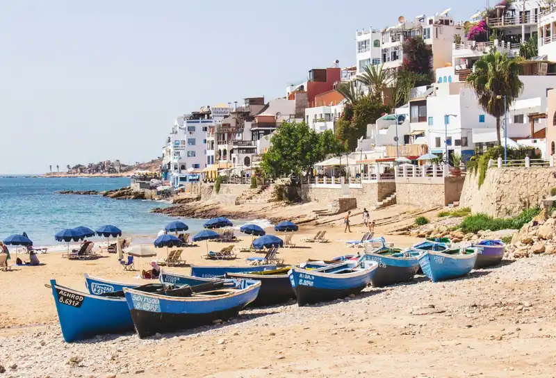 Blue wooden fishing boats pulled up on the beach below the white houses of Taghazout